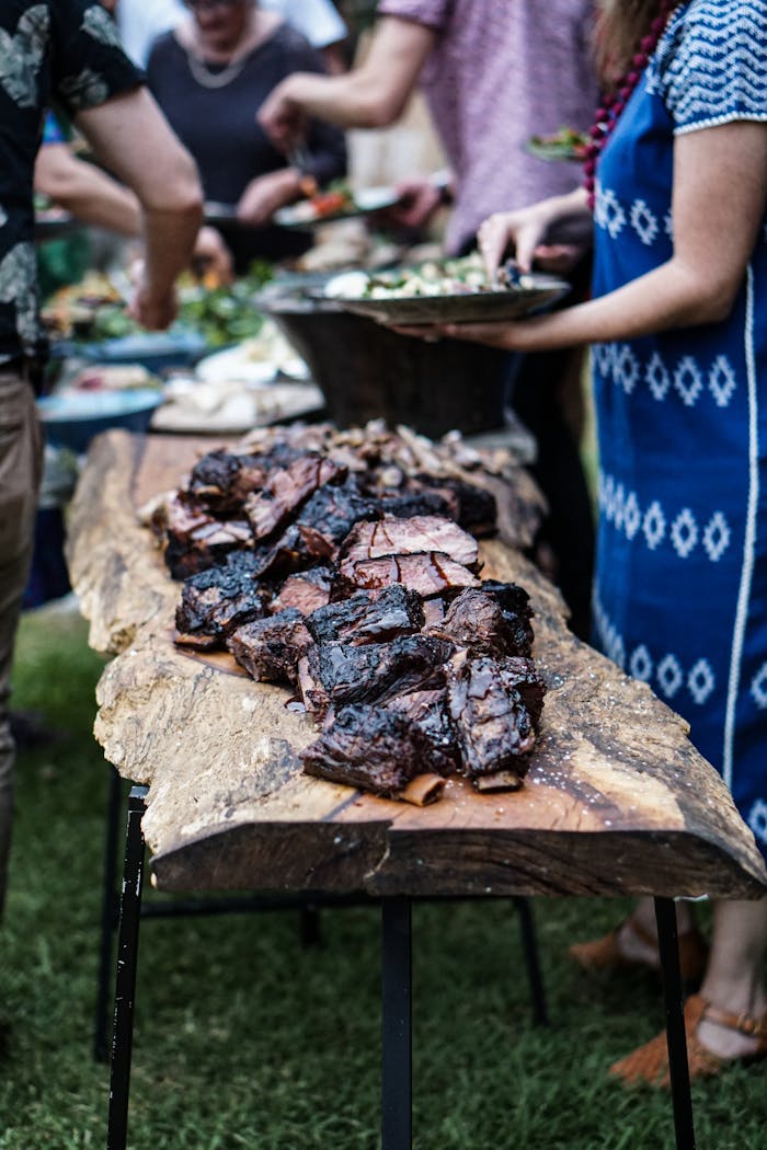 A group of people enjoying a summer barbecue with grilled meat on a wooden table outdoors.